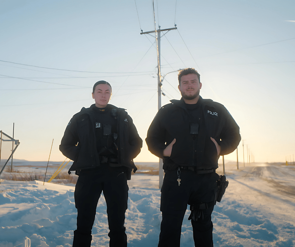 Two police officers stand in the snow on a rural Northwestern Ontario road at sunset, with power lines and open fields in the background. Both wear dark uniforms and jackets, looking at the camera—guardians of safety in this remote region.