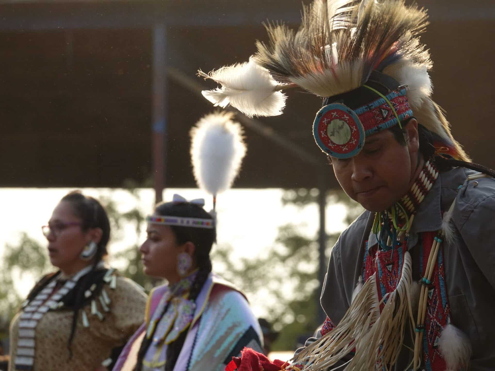 Three dancers in traditional First Nations regalia at a pow wow. The dancer in front wears an elaborate roach headdress with feathers and beadwork.
