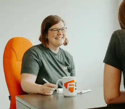 A woman wearing glasses and a grey t-shirt sits at a desk, smiling and holding a pen. She has a white mug with an orange logo, suggesting a meeting about Vendor of Record procedures with another person seated opposite her.