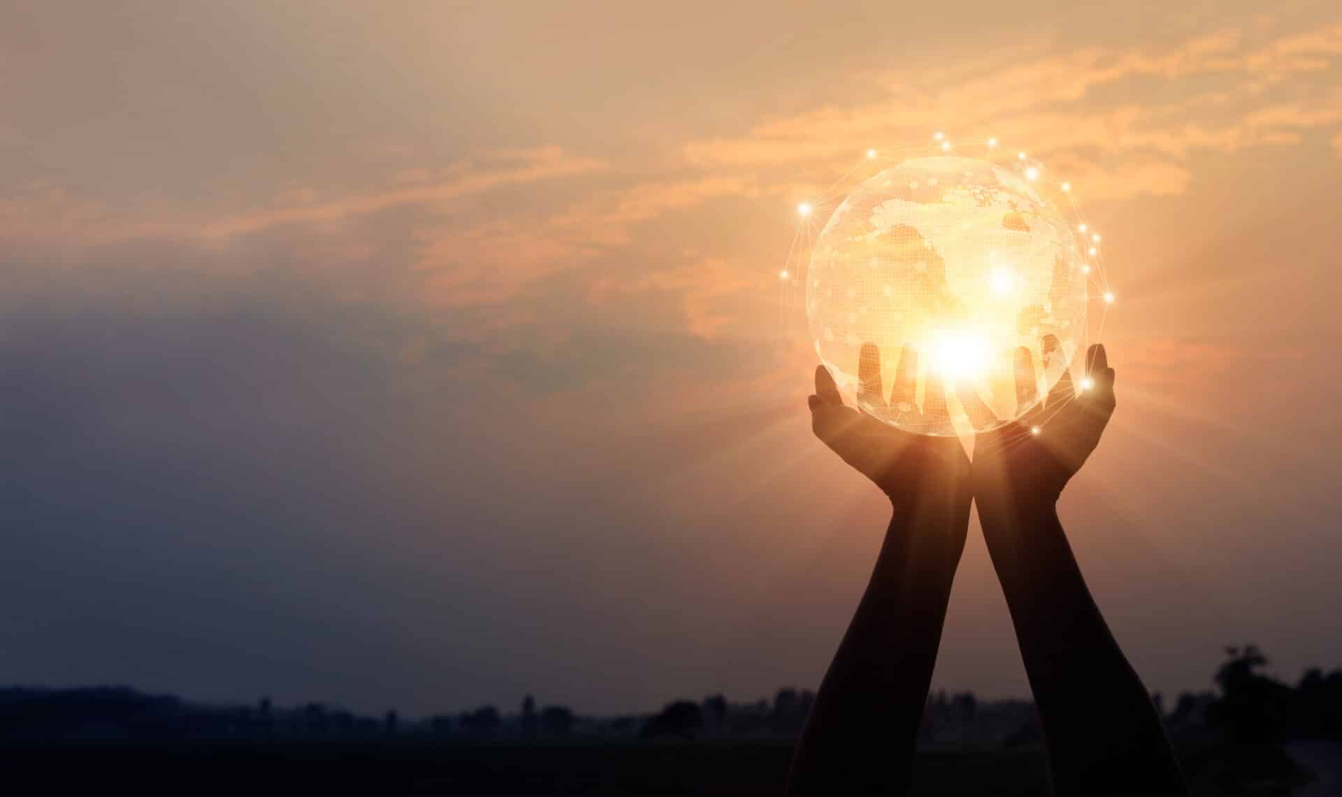 Two hands hold up a glowing digital globe against a sunset sky, with light points highlighting global connections. The scene symbolises unity, hope, and global connectivity.
