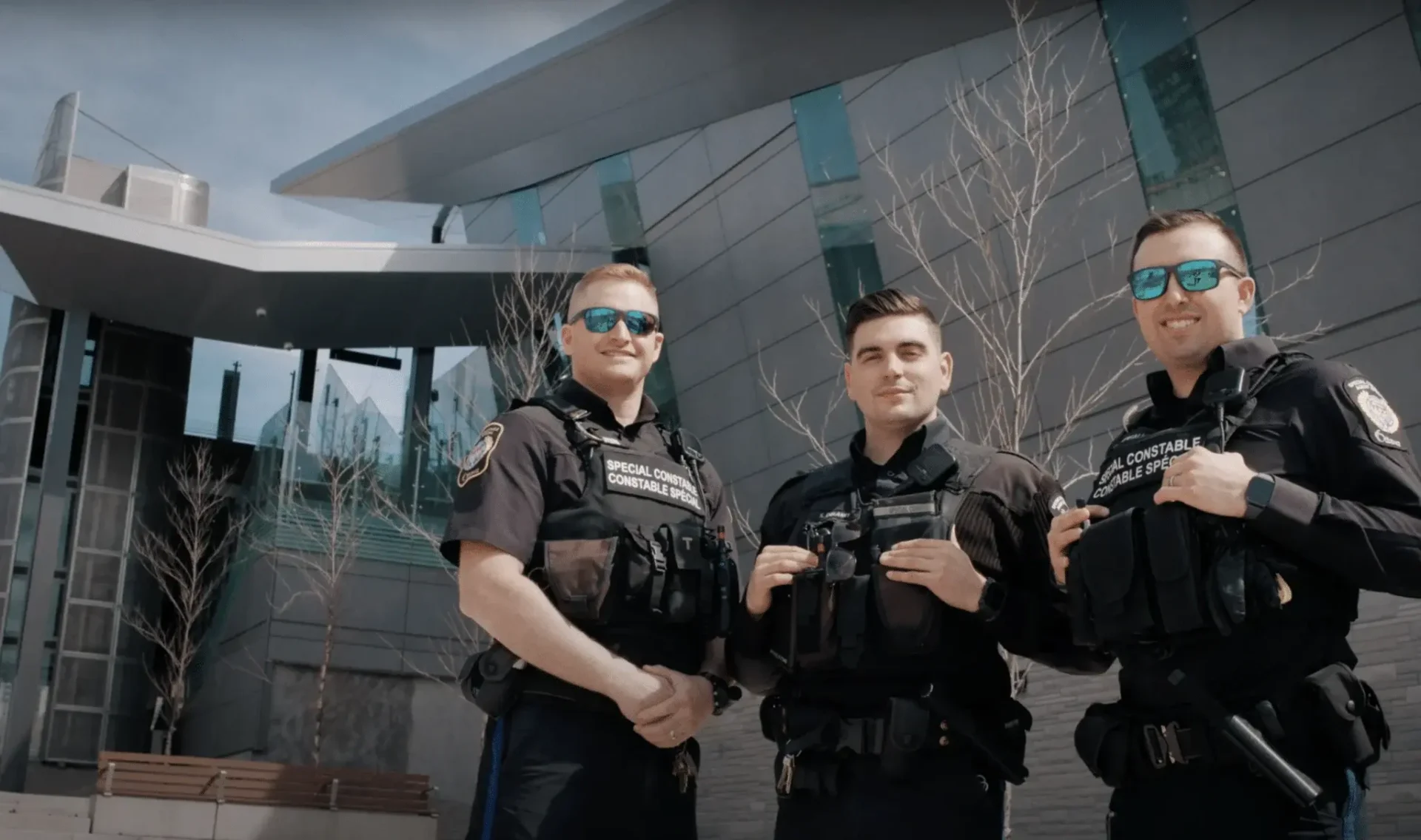 Three uniformed police officers stand outside a modern building with large windows, smiling and wearing sunglasses—showcasing the positive side of law enforcement technology within the municipal government. Leafless trees are visible in the background.
