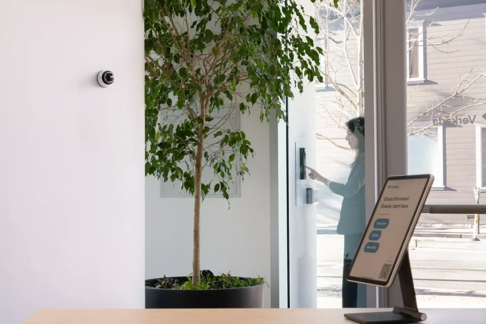 A security camera on a white wall faces a large potted plant near a glass door, where a person uses a touchscreen panel—highlighting the importance of cyber security in municipal government buildings. A digital check-in screen sits on the wooden desk in the foreground.