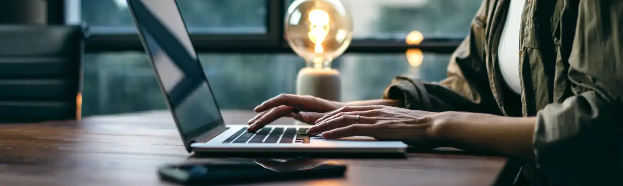 Person typing on a laptop at a wooden desk with a vintage Edison bulb glowing in the background